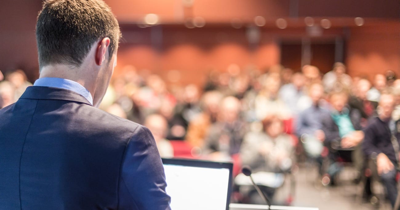 Behind image of a man giving a presentation to a whole room of people