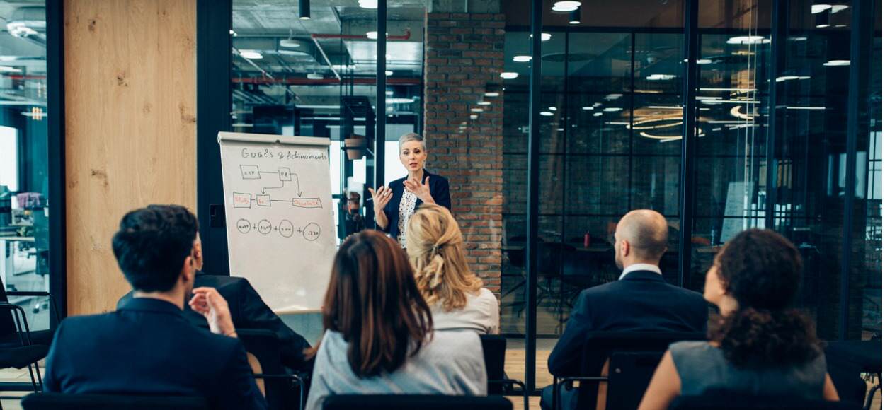 Female employee in black suit jacket leading team meeting in glass office next to a whiteboard