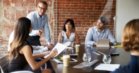 Older Boss Leading Staff Meeting In Modern Boardroom Through Glass