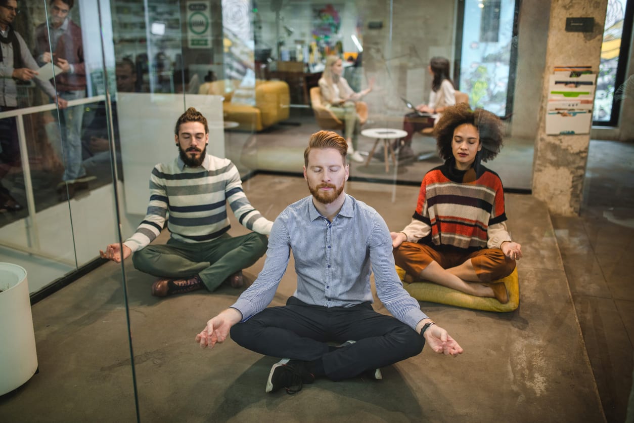 Young business team exercising Yoga in Lotus position on the floor at casual office. The view is through glass wall.