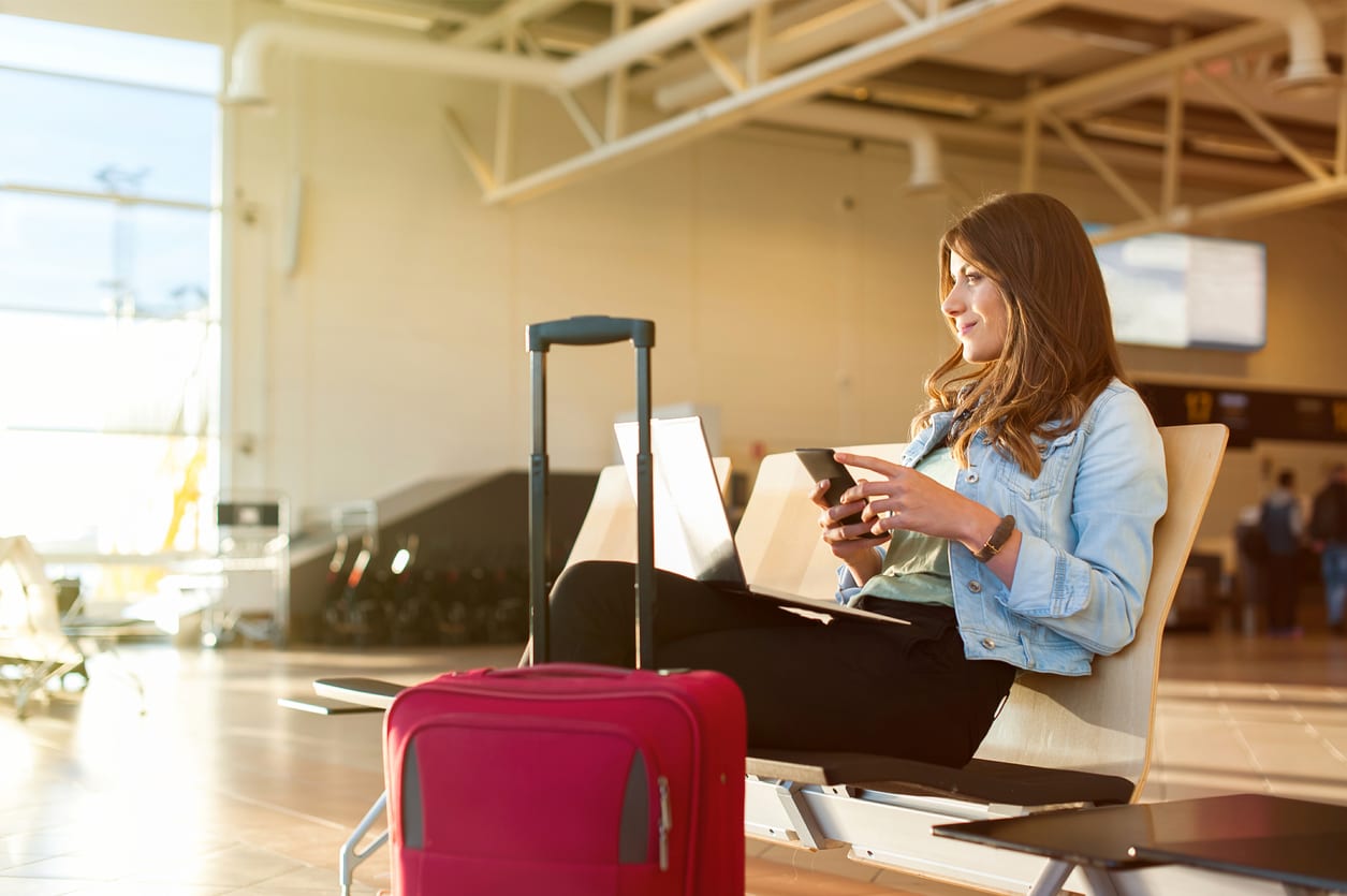 Airport Young female passenger using smartphone and laptop sitting in terminal hall while waiting for her flight