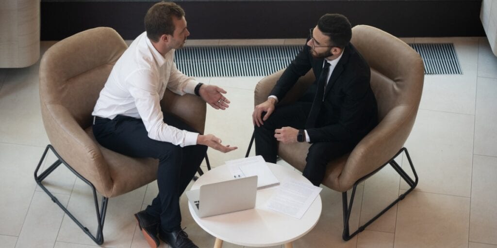 Two smartly dressed male employees sat in arm chairs discussing something in front of a laptop screen on the table