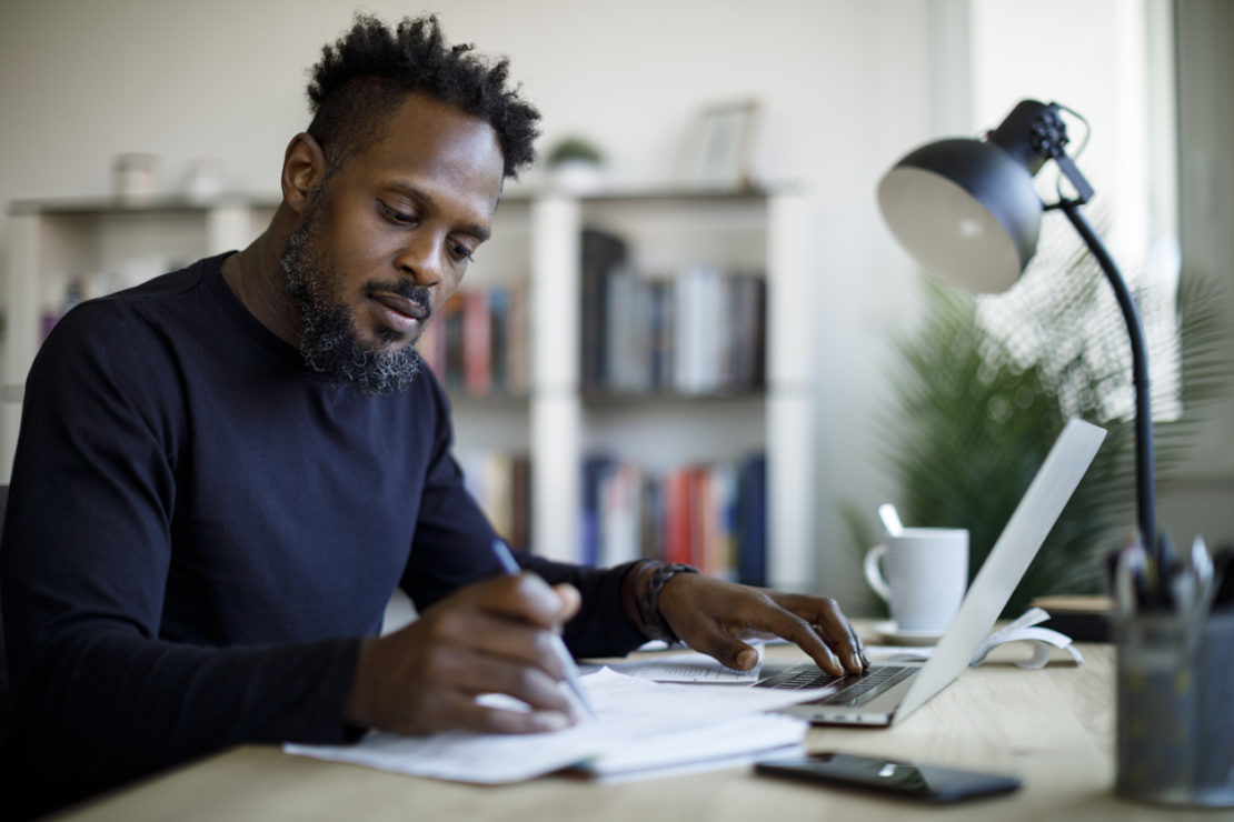 Dark skinned male in navy turtleneck jumper working at a desk whilst on laptop and writing notes on a notebook