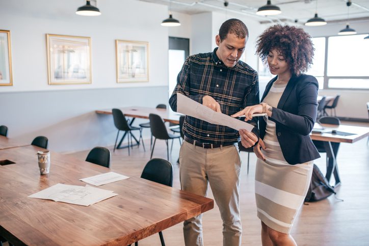 Man and women stood up in empty room both looking and pointing at a big piece of paper