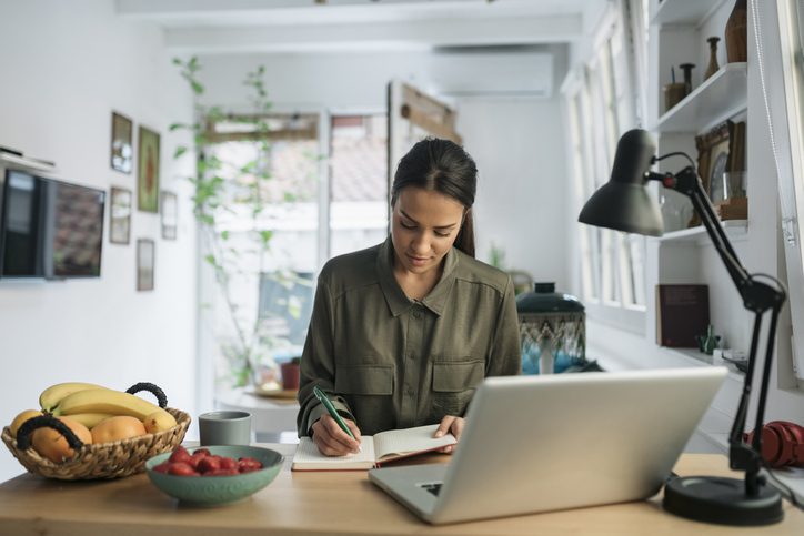 Young women writing in a notepad in her living room in front of a laptop