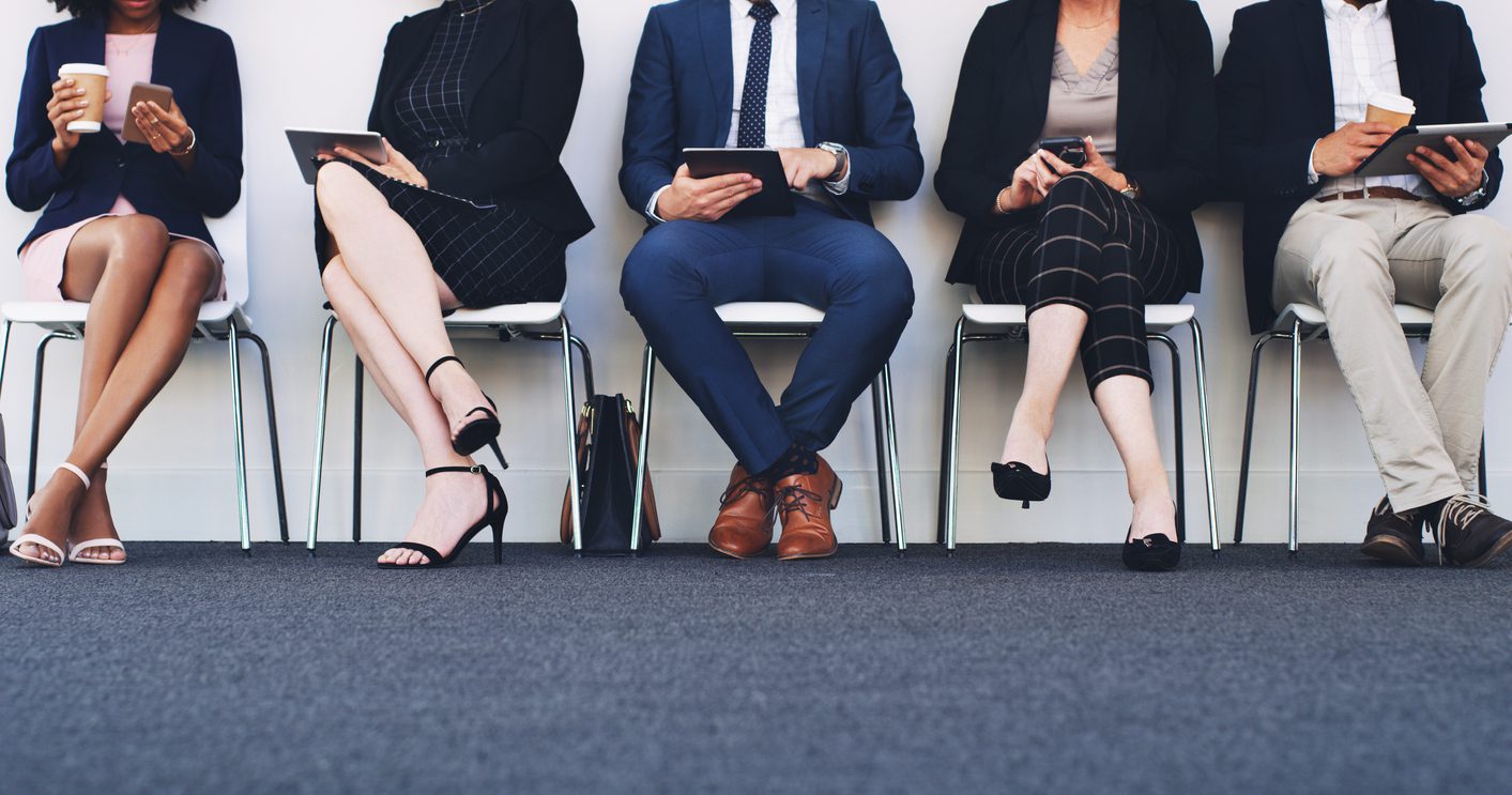 Cropped shot of a diverse group of businesspeople using technology while sitting in a line in the office waiting for an interview