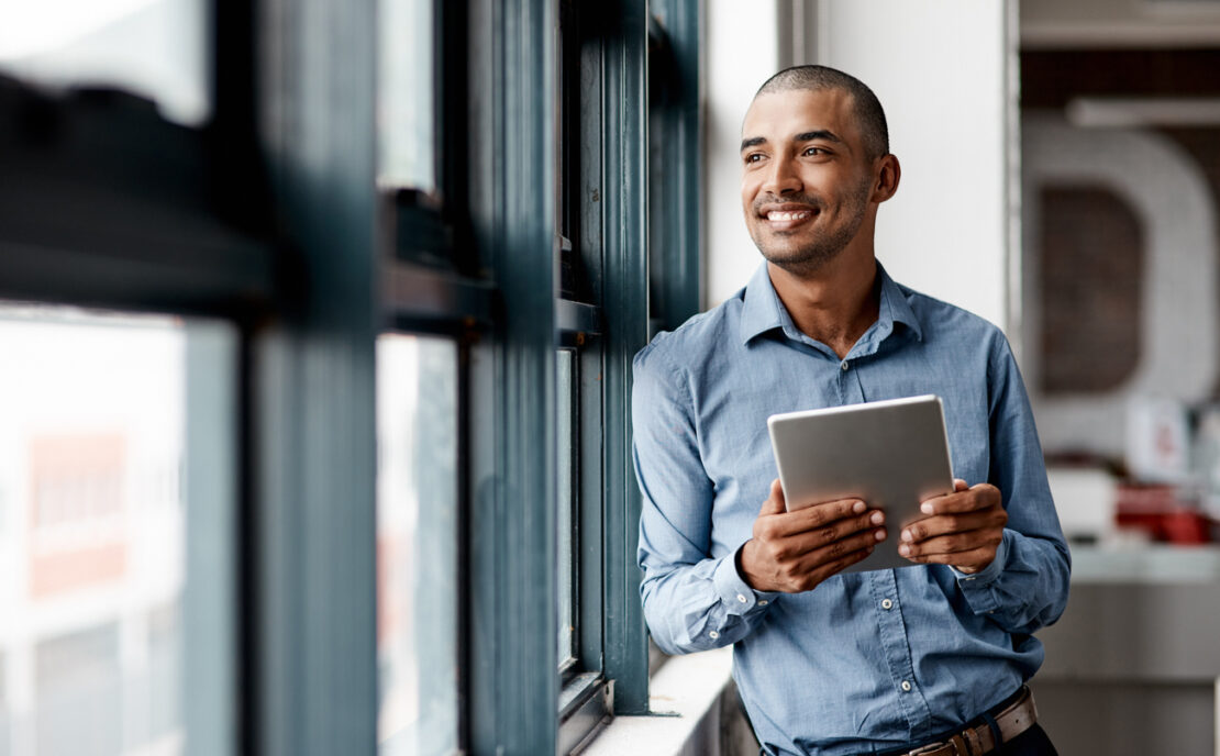 Smartly dressed male employee smiling looking out the window whilst holding a tablet