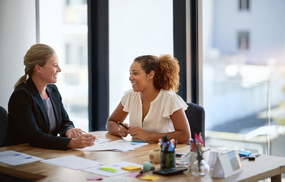 Two females smiling and talking on a desk within the office