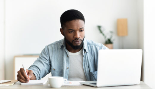 Dark skinned male focused on laptop screen whilst taking notes