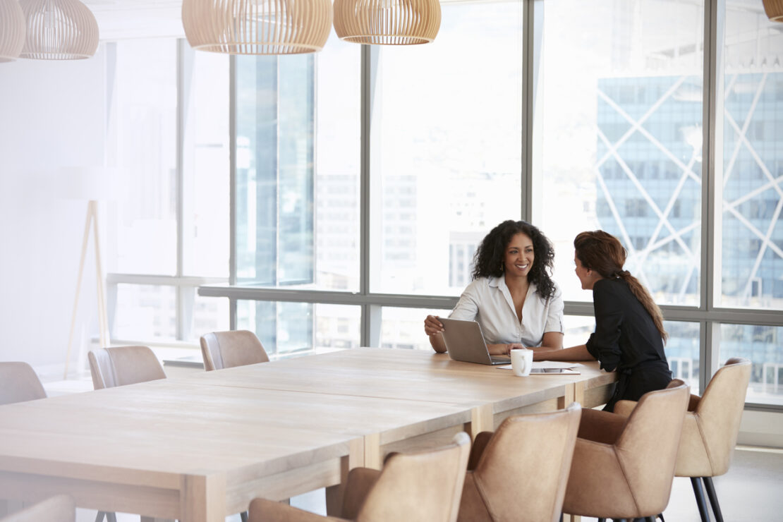 Zoomed out image of two females speaking in a large empty meeting room