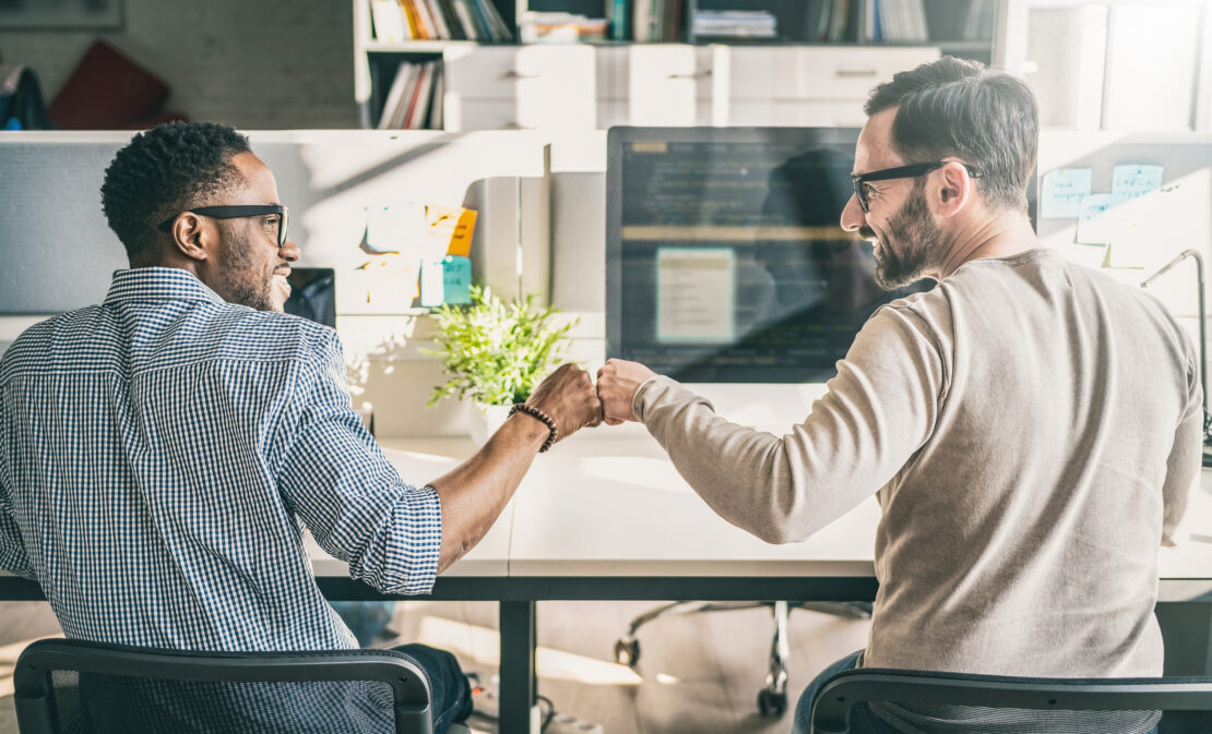 Two male employees fist bumping at their desks