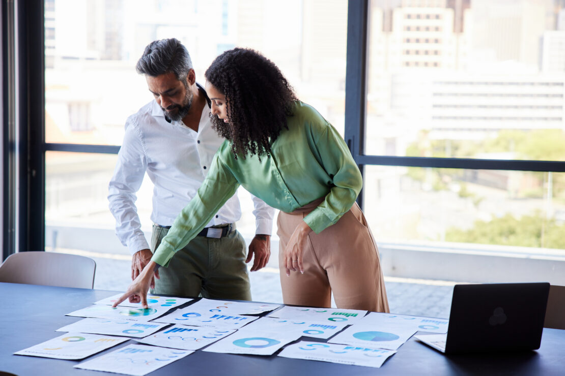 Two employees stood up over meeting table pointing at data charts on a table